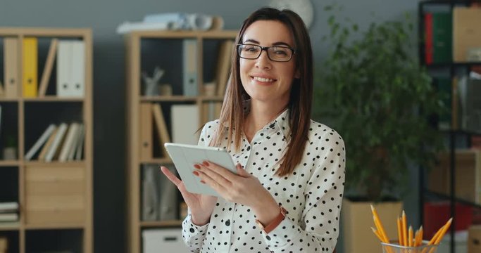 Young attractive Caucasian businesswoman standing in the office and using tablet device, then rising her head and smiling to the camera happily. Portrait shot.