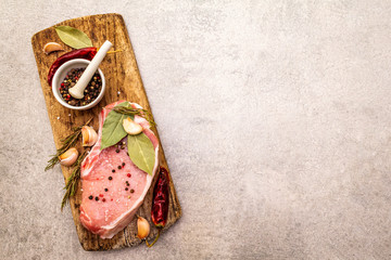 Raw pork steak with spices and dried herbs on vintage wooden board. Salt, garlic, hot pepper, rosemary, bay leaf with ceramic mortar and pestle on a stone background, top view.