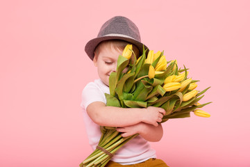 Adorable smiling child with spring flower bouquet looking at camera isolated on pink. Little toddler boy in hat holding yellow tulips as gift for mom. Copy space for text 
