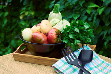 Fresh garden vegetables and herbs: tomatoes, potatoes, onion, parsley, cilantro, zucchini assorted in big black glass bowl  on wooden table with kitchen stuff on nature background, closeup. 