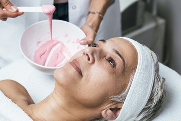 Beautiful senior woman receiving facial mask with rejuvenating effects in spa beauty salon.