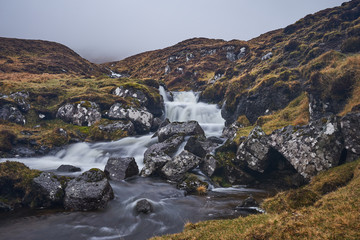 Fototapeta na wymiar Wild stream or creek in moorland or heath in the highland of Faroe island Vagar with waterfall cascades and stones with moss and lichen on them. Picture taken in cloudy, misty, foggy and rainy day.