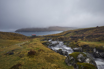Landscape Picture of countryside in Faroe islands shown sheep grazing or pasture land in moorland on top of hill in fjord, wired fences, wild creek with water cascade during typical spring rainy day.