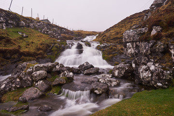 Wild creek or stream with waterfalls and water cascade between fences of sheep pasture or grazing land during the cold, foggy, místy and rainy day in spring on Faroe islands in north atlantic ocean.