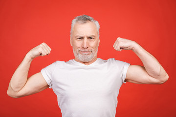 Check this out! Senior man showing muscle. Aged man flexing his arms isolated against red background. Cheerful excited modern cool pensioner grandpa practising bodybuilding.