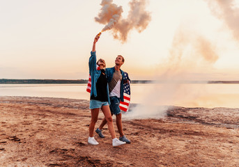 american couple walking along the beach and holding hand flare or fusee covered in american flag. Freedom and independence concept