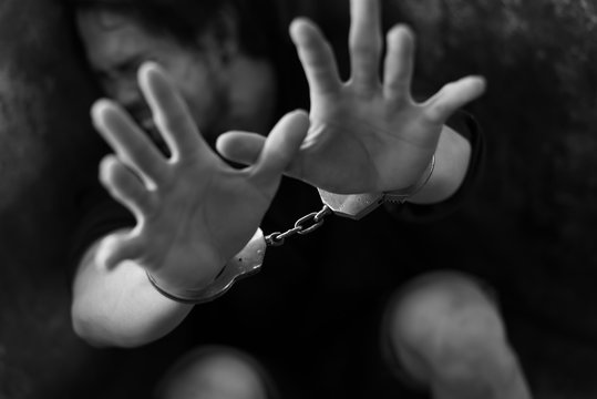 Man Prisoner With His Hands Shackled In Chains On A Dark Background.