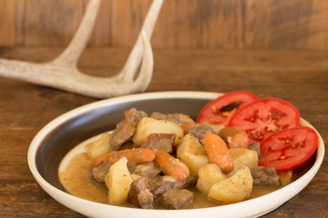 Venison Stew with Tomato Slices on Rustic Table with Deer Antlers in Background