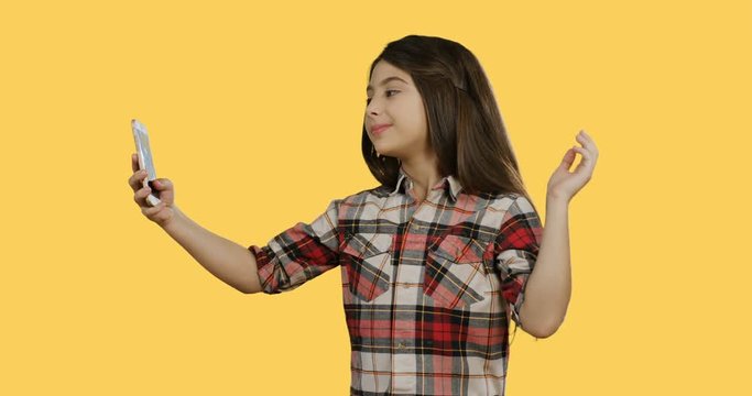 Caucasian teen small girl in a plaid blouse making selfies on the smartphone and posing while standing on the yellow screen background.