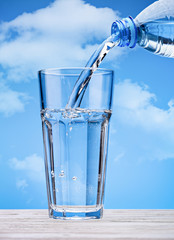 Pouring drinking water from plastic bottle into glass, against  blue sky with clouds. Sparkling water with bubbles in large glass. Copy space.