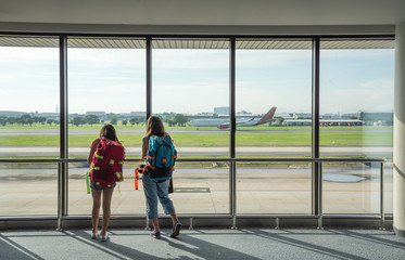Two young women backpacking, waiting for plane at the airport to travel on holiday. Back side of traveler girl looking at the flying plane. travel and active lifestyle concept