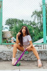 Summer lifestyle image of trendy pretty young girl sitting next to the skateboard coart with her plastic skateboard.