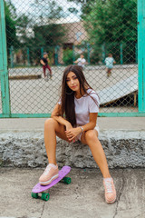 Summer lifestyle image of trendy pretty young girl sitting next to the skateboard coart with her plastic skateboard.