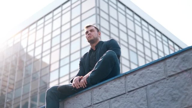 low angle shot handsome man sits on black parapet against modern building reflecting sunlight