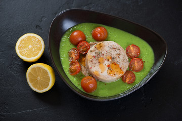 Black bowl with baked fish medallions, green peas puree, cherry tomatoes and lemon. Studio shot on a black stone background