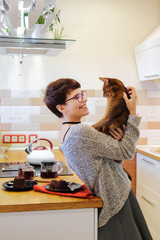 Smiling woman playing with a ginger cat, hugging and fooling around in the kitchen with him.