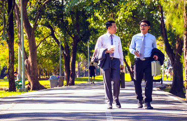 Businessman, They are walking on road in park. They are talking  business. He is drinking coffee and His friend holding business bag. Photo concept business and relax time.