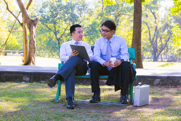 Fototapeta premium Businessman, They are sitting on bench in park. He is play notebook and search internet. Another one holding paper and talking about business in relax time. Photo concept business and relax time.