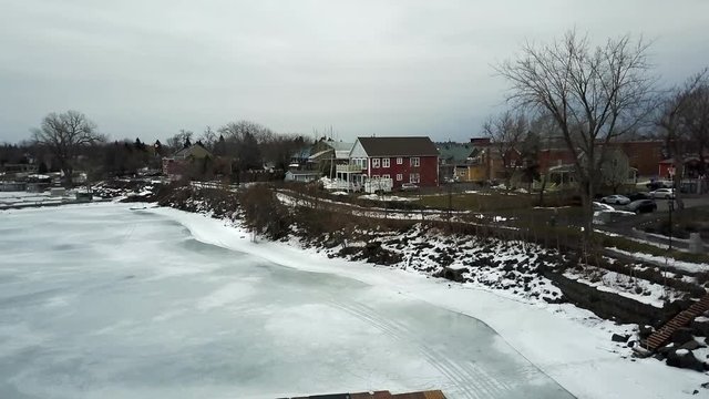 Cinematic aerial / drone footage descending over the lake with dry trees, snow, houses, a small town, cars in the background in Chambly, Quebec, Canada during winter season.