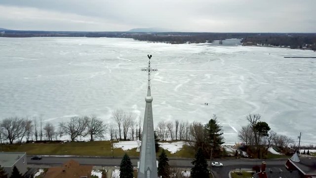 Cinematic aerial / drone footage rolling up the camera towards a church, with a religious cross, frozen lake, houses, cars, mountains in the background in Chambly, Quebec, Canada during winter season.