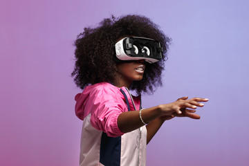 Mod young brown-haired curly girl dressed in the pink sports jacket uses the virtual reality glasses in the studio on neon background