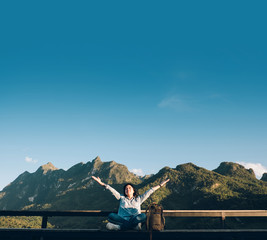 Asian woman traveler  sitting and arms up in the air at view point terrace at landscape view of mountain with cloud and blue sky in sunny day at forest.backpacker explore nature life.