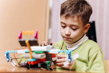 Diligent boy dressed in green sweater sits at the desk with computer and makes the robot from the robotic constructor in the School of Robotics