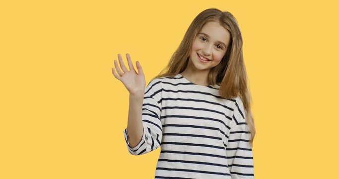 Close up of the blonde teenager girl with long hair in the striped blouse waving her hand and smiling on the yellow wall background. Portrait.