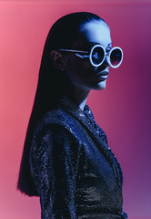 Fashion girl with long hair and round sunglasses in a black shining dress poses in neon light in the studio