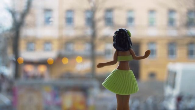 Dancing Hawaian plastic hula girl doll looking out a window onto an urban street with blurred traffic and pedestrians rushing by