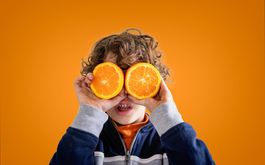 Cute kid playing with orange fruit covering his eyes with half piece of the fruit over a vivid orange background