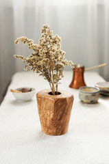A wooden vase made of oak burl with dry yellow flowers standing on a white table cloth. Coffee pot with cups and a little bowl with chocolate are on the background.
