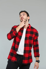 Cute young man with a beard in a red-black checkered shirt holds a hand over his face, isolated on a gray background.