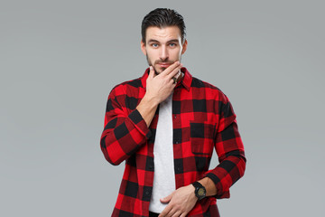 Handsome young man with a beard in a red-black checkered shirt holding a hand over his face, isolated on a gray background.
