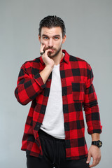 Handsome young man with a beard in a red-black checkered shirt holding a hand over his face, isolated on a gray background.