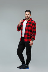 Handsome young man with a beard in a red-black checkered shirt standing tall, isolated on a gray background.