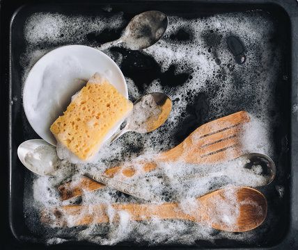 A white plate, a knife, a sponge, wooden kitchen spatulas and spoons in the detergent foam on a black oven-tray. Washing dishes concept. Top view.