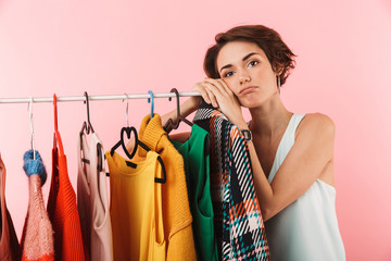 Woman stylist posing isolated over pink wall background near a lot of clothes.