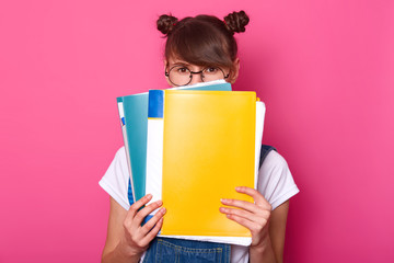 Image of surprised emotive european woman hides behind colourful paper folders, wears round black glasses, white t shirt, denim overalls, stands against pink background. Back to school concept.