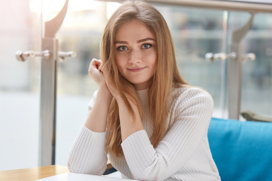 Headshot of pleasant looking woman with pleased facial expression, looks at camera, dressed in casual clothes, sits on confortable sofa in cafeteria, waits for order, spends free time in public place
