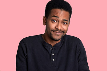 African man with beard and mustache crews his eyes and smiles, male looks shy. Model stands isolated over pink blackground, poses and looks at camera. Young guy wears black shirt. People concept.
