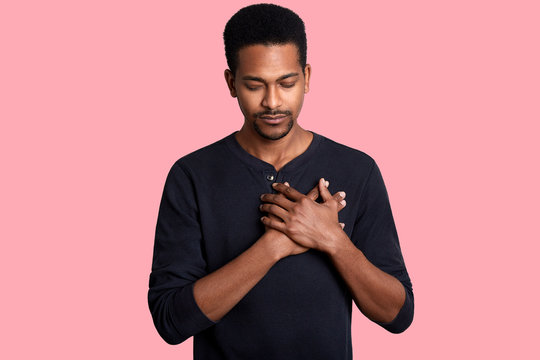 Grateful calm african man holds hands on chest, feels love, gratitude, honesty. Dark skinned guy thanks isolated on pink studio background. Female with closed eyes dresses black casual shirt.
