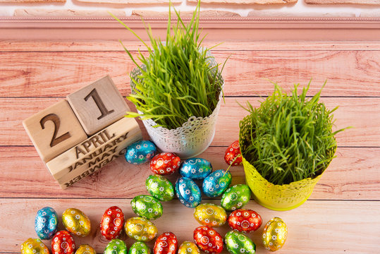 Top view on Easter composition. Variety of small different colors eggs in foil laying on table near wooden cube calendar and flower pots with green grass stems