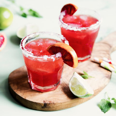 Blood orange and lime margarita cocktail with slices of fresh fruit and ice on optimistic green background.