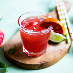 Blood orange and lime margarita cocktail with slices of fresh fruit and ice on optimistic green background.