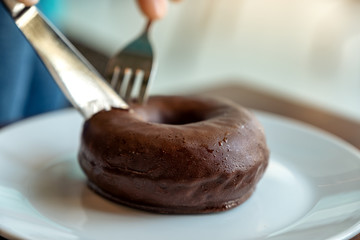 Closeup image of hands cutting a piece of chocolate donut by knife and fork for breakfast on wooden table