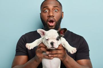 Dog ownership concept. Stupefied man carries in front white puppy with black ear, shocked to hear...