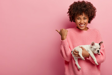 Happy dark skinned girlfriend shows way to animal shop, feels satisfied with good consultancy in vet clinic, carries french bulldog, indicates away with thumb, isolated over pink background.