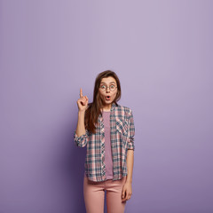 Impressed female shop assistant looks up amazed, points with fore finger, says wow, wears casual clothes, suggests to look at awesome product, advertises something, isolated over purple wall.