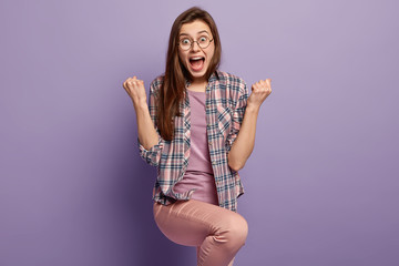 Overjoyed lady raises fists and knee in triumph, exclaims with happiness, celebrates entering...